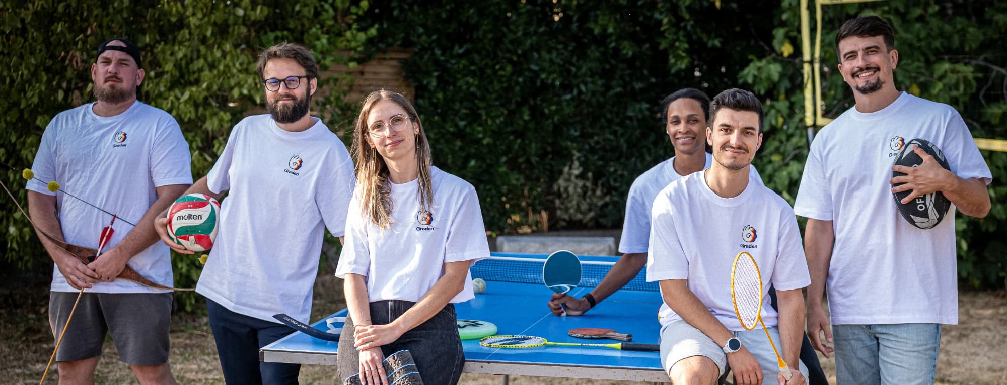 L’équipe Graden, sept personnes en t-shirt Graden autour d’une table de ping-pong, avec un arc, un ballon de volley, un ballon de rugby et des raquettes.